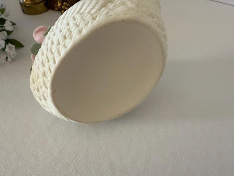 May include: Off-white ceramic swan-shaped container with a textured exterior. The container has a basket weave pattern and is decorated with pink and green floral accents. The bottom of the container is smooth and unadorned.
