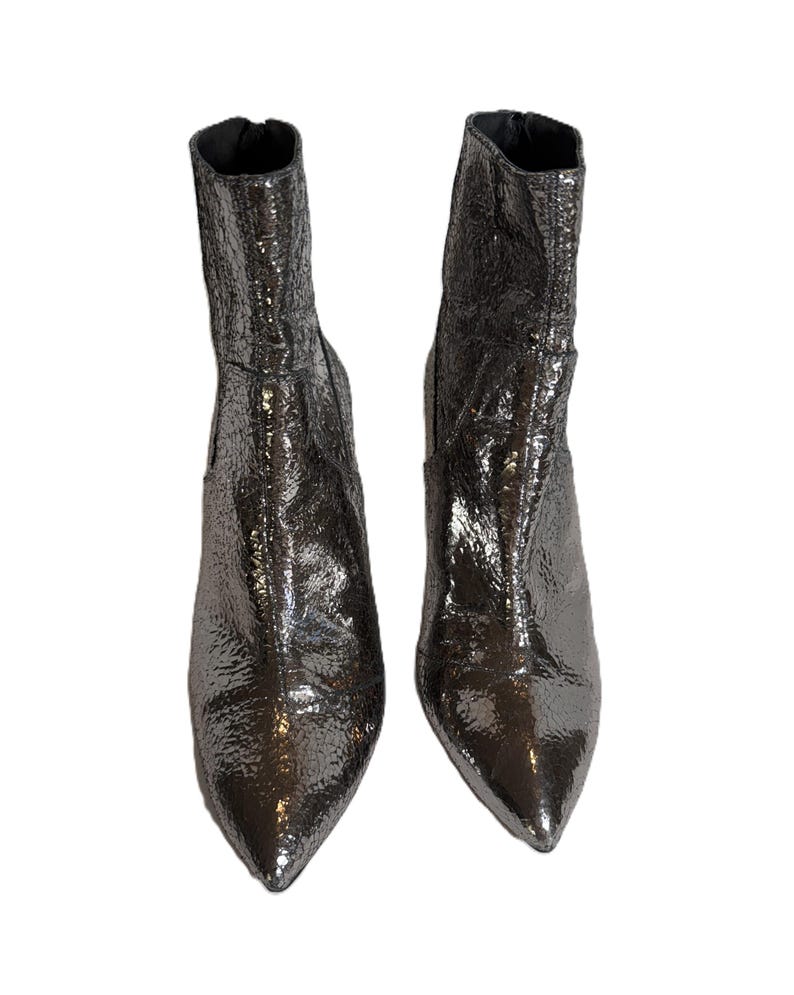 May include: A pair of metallic silver ankle boots with pointed toes. The boots have a textured, crinkled appearance and a small heel. The boots are displayed against a white background.