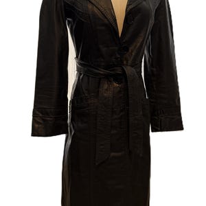 May include: A black leather trench coat with a belted waist and button closure. The coat features a notched collar, long sleeves, and two front pockets. The coat is displayed on a mannequin.