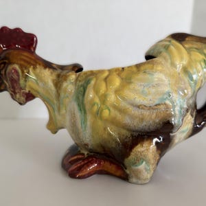 May include: A ceramic rooster-shaped pitcher with a handle. The pitcher features a red comb, brown, yellow, and green mottled glaze. The rooster's feet are a deep red-brown color.