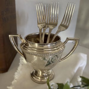 May include: A polished silver-toned sugar bowl with two handles and a decorative beaded rim, containing four matching silver-toned forks. The bowl features a tapered body resting on a circular pedestal base with a subtle floral design on the front.