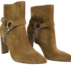 May include: A pair of brown suede ankle boots with pointed toes and block heels. Each boot features a strap with a silver-tone buckle and a decorative chain. The boots have a stacked heel.