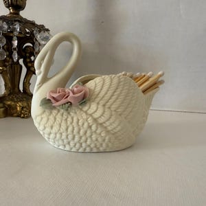 May include: A cream-colored swan-shaped match holder with a textured body and a curved neck. Two pink roses with green leaves adorn the side. The holder is filled with wooden matches. A decorative lamp is in the background.