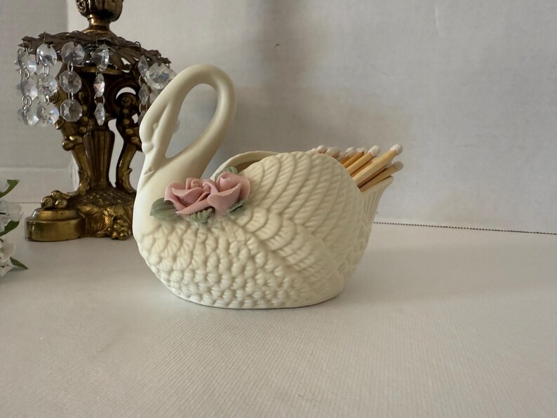 May include: A cream-colored swan-shaped match holder with a textured body and a curved neck. Two pink roses with green leaves adorn the side. The holder is filled with wooden matches. A decorative lamp is in the background.
