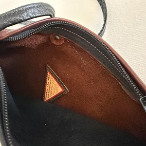 Puede incluir: Vista abierta de un bolso de hombro de cuero negro y marr&oacute;n. El interior es marr&oacute;n con una base negra. Es visible una peque&ntilde;a etiqueta triangular con texto. El bolso tiene una cremallera y una correa negras.