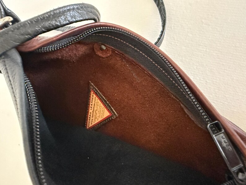 Puede incluir: Vista abierta de un bolso de hombro de cuero negro y marr&oacute;n. El interior es marr&oacute;n con una base negra. Es visible una peque&ntilde;a etiqueta triangular con texto. El bolso tiene una cremallera y una correa negras.