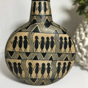 Puede incluir: Un jarrón redondo de color beige con cuello estrecho, decorado con patrones negros pintados a mano. El diseño incluye filas de figuras estilizadas tomadas de la mano, intercaladas con bandas geométricas de triángulos y líneas verticales.