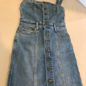 Peut inclure: Une robe en jean bleu avec un devant boutonn&eacute; et des bretelles fines. La robe a un corsage ajust&eacute; et une jupe l&eacute;g&egrave;rement &eacute;vas&eacute;e.
