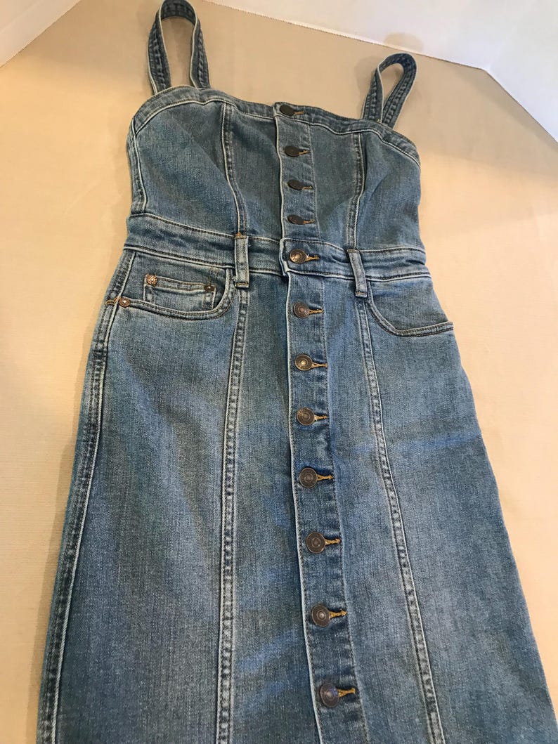 Peut inclure: Une robe en jean bleu avec un devant boutonn&eacute; et des bretelles fines. La robe a un corsage ajust&eacute; et une jupe l&eacute;g&egrave;rement &eacute;vas&eacute;e.