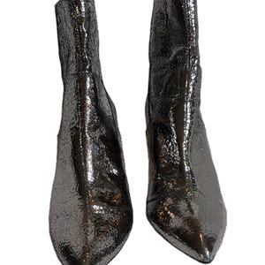 May include: Two metallic silver ankle boots with a textured, crackled finish. The boots feature pointed toes and a mid-height heel. The shiny material reflects light, creating a dynamic appearance. The boots are set against a white background.