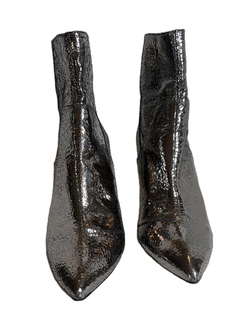 May include: Two metallic silver ankle boots with a textured, crackled finish. The boots feature pointed toes and a mid-height heel. The shiny material reflects light, creating a dynamic appearance. The boots are set against a white background.