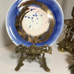 May include: A white ceramic plate with a blue and gold design. The plate has a circular design with a gold center and blue rings around it. The plate is on a gold metal stand.