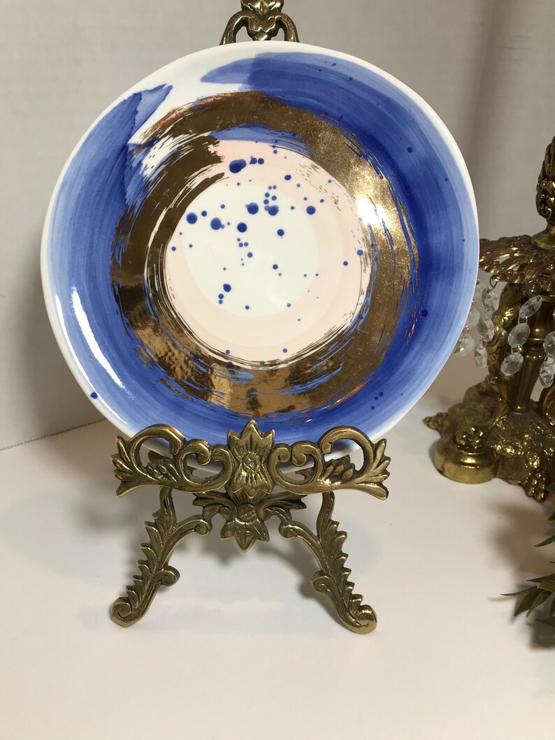 May include: A white ceramic plate with a blue and gold design. The plate has a circular design with a gold center and blue rings around it. The plate is on a gold metal stand.