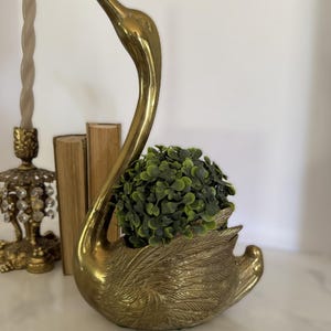 Puede incluir: Una figura de cisne de latón pulido con texturas de plumas detalladas en el cuerpo y un cuello largo y curvado. El centro hueco del cisne contiene una pequeña planta verde artificial redonda con numerosas hojas diminutas.