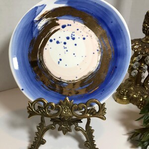 May include: A white ceramic plate with a blue and gold abstract design. The plate is on a gold metal stand.