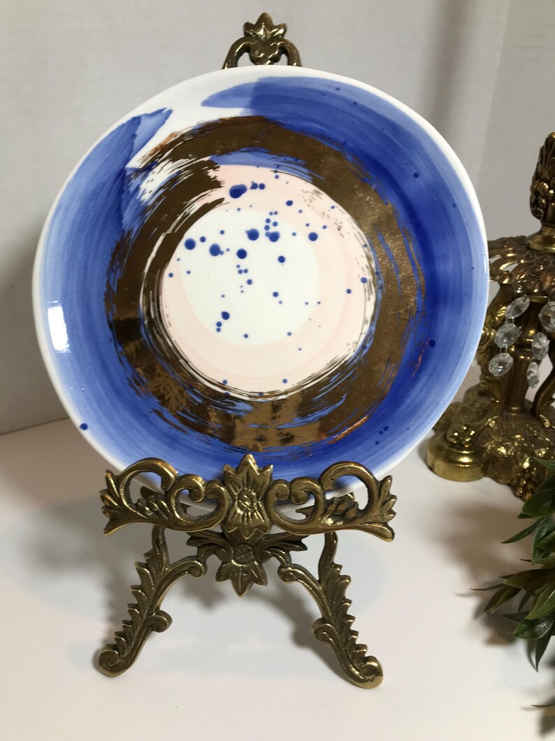 May include: A white ceramic plate with a blue and gold abstract design. The plate is on a gold metal stand.