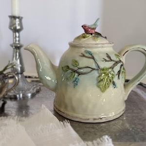 May include: A cream-colored ceramic teapot featuring a raised relief design of brown branches with green leaves and blue buds. The lid is topped with a small, sculpted pink and blue bird. The teapot has a curved handle and a tapered spout.