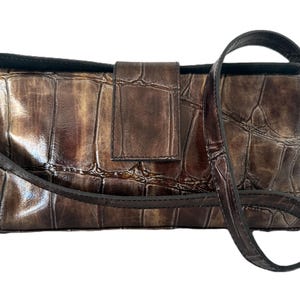 May include: A brown, rectangular handbag with a textured, crocodile-like pattern. The bag features a flap closure and a long, thin shoulder strap. The color is a mix of brown and bronze.