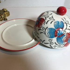 May include: A white ceramic serving dish with a red rim and a matching domed lid. The lid is decorated with blue and red floral designs and a red knob. The dish is likely used for serving food.