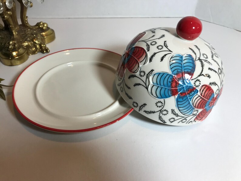 May include: A white ceramic serving dish with a red rim and a matching domed lid. The lid is decorated with blue and red floral designs and a red knob. The dish is likely used for serving food.