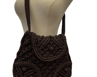 Könnte beinhalten: Eine braune Makramee-Umhängetasche mit gewebtem Design. Die Tasche hat einen Klappenverschluss und einen langen, geflochtenen Riemen. Die Tasche wird auf einer Schaufensterpuppe präsentiert.