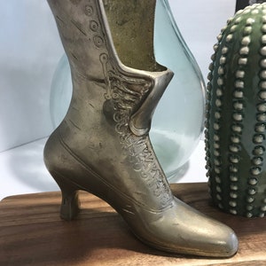 May include: A silver metal boot shaped vase with a high heel. The boot is sitting on a wooden surface.