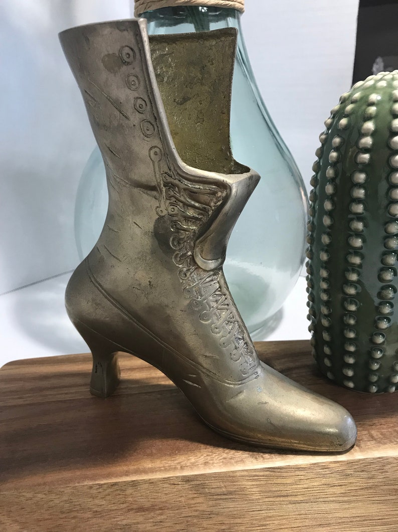 May include: A silver metal boot shaped vase with a high heel. The boot is sitting on a wooden surface.