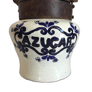 May include: A white ceramic jar with a dark brown metal lid and handle. The jar is decorated with blue floral designs and the word "AZUCAR" in blue script. The jar has a crackled glaze finish, giving it a vintage appearance.