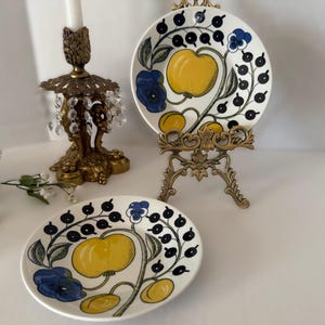 May include: Two round ceramic plates featuring a botanical illustration of a large yellow fruit, smaller yellow fruits, blue flowers, and black berries on a white background. One plate rests on a gold metal stand, and one lies flat.