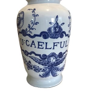 Könnte beinhalten: Eine weiße Keramikvase mit blau handgemalten Illustrationen. Die Vase zeigt einen Vogel, florale Elemente und einen Obstkorb. Das Wort "U:CAELFUL" ist in Blau geschrieben. Die Vase hat einen breiten Körper und einen schmalen Hals.