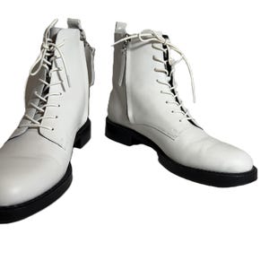 May include: A pair of white leather boots with black soles. The boots have laces and side zips. The boots are a classic style, suitable for various outfits.