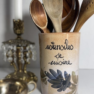 May include: A ceramic utensil holder with the words "ustensiles de cuisine" in black script. The holder is beige with a floral design in blue and brown. Wooden spoons of various shapes and sizes are inside.