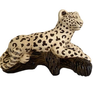 May include: A ceramic leopard figurine in a resting pose, featuring a cream-colored body with dark brown spots. The leopard rests on a dark brown, textured log, creating a naturalistic scene. The figurine is a decorative item.