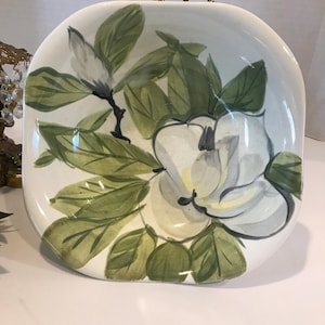 May include: A white ceramic bowl with a hand-painted design of a magnolia flower and leaves. The flower is white with a yellow center and the leaves are green.
