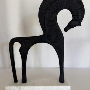 May include: A black, stylized horse sculpture with an arched neck and intricate carved details. The horse stands on a white marble base. The sculpture's design features geometric patterns and a circular emblem.