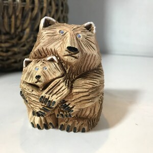 May include: A carved wooden bear figurine showing a mother bear embracing a cub. The bears are light brown with textured fur details, black noses and paws, and blue eyes. The figurine is a decorative item.