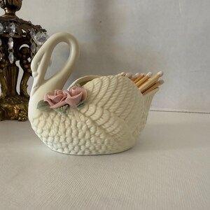 May include: Cream-colored swan-shaped match holder with a textured body and a curved neck. Two pink roses with green leaves adorn the side. The holder is filled with wooden matches.