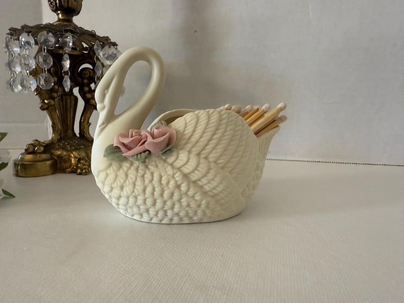 May include: Cream-colored swan-shaped match holder with a textured body and a curved neck. Two pink roses with green leaves adorn the side. The holder is filled with wooden matches.