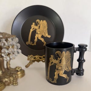 May include: A black ceramic mug and plate set with a gold illustration of a figure with wings. The mug has a unique handle design. The plate is round and the mug is cylindrical. The set is displayed on a gold stand.
