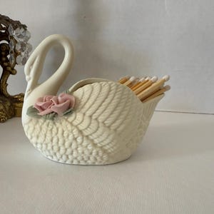 May include: A cream-colored ceramic swan match holder with a pink rose accent. The swan's wings are detailed with a textured pattern, and the holder is filled with wooden matches. A decorative lamp is in the background.
