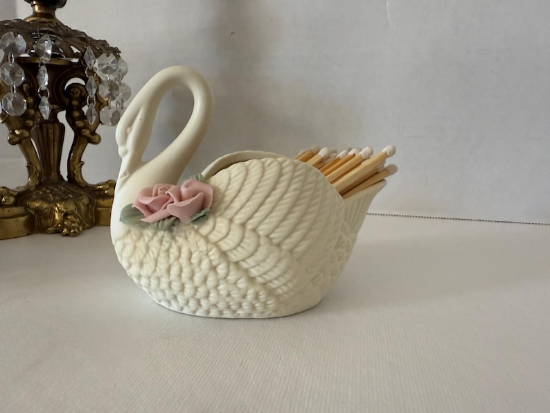 May include: A cream-colored ceramic swan match holder with a pink rose accent. The swan's wings are detailed with a textured pattern, and the holder is filled with wooden matches. A decorative lamp is in the background.