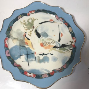 May include: A white ceramic plate with a blue rim and a gold trim. The plate features a watercolour painting of a bird with a red, yellow, and green design. The plate also has a black and white design in the centre with the word "生" in black.