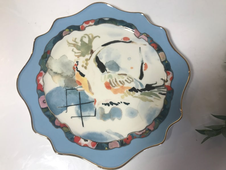 May include: A white ceramic plate with a blue rim and a gold trim. The plate features a watercolour painting of a bird with a red, yellow, and green design. The plate also has a black and white design in the centre with the word "生" in black.