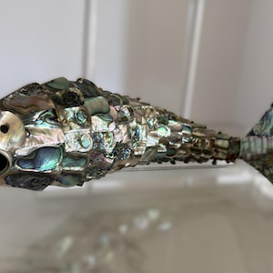 May include: A decorative fish figurine covered in iridescent abalone shell tiles. The surface features a mosaic of shimmering green, blue, and purple hues. The fish has an open mouth, a distinct eye, and a wide, fan-shaped tail fin, all crafted from polished shell fragments.