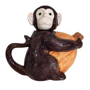 May include: A ceramic monkey teapot with a brown body, a white face, and a tan banana. The monkey is holding the banana. The teapot has a curved handle and a spout.