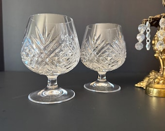 May include: Two clear crystal brandy glasses with detailed cut glass patterns. The glasses have a rounded bowl, a short stem, and a wide base. The cut glass design features diamond and leaf-like patterns. The glasses are displayed on a dark surface.