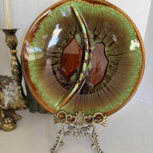 May include: A round ceramic dish featuring a glossy glaze with a vibrant green and brown drip pattern. The dish is divided into two sections by a central curved ridge and is displayed on an ornate, metallic, gold-toned decorative stand.