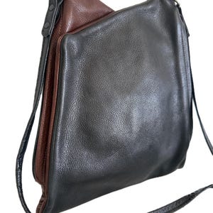 Puede incluir: Bolso de hombro de cuero bicolor en negro y marr&oacute;n. El bolso tiene forma rectangular con una correa larga y fina. El cuero tiene un aspecto texturizado y el bolso presenta herrajes plateados.
