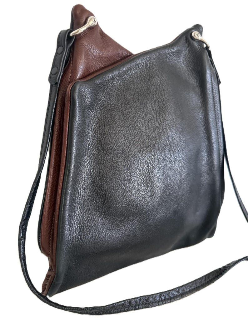 Puede incluir: Bolso de hombro de cuero bicolor en negro y marr&oacute;n. El bolso tiene forma rectangular con una correa larga y fina. El cuero tiene un aspecto texturizado y el bolso presenta herrajes plateados.