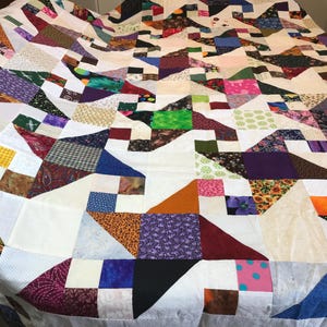 Puede incluir: Un edredón hecho a mano con un diseño de patchwork. El edredón presenta una variedad de telas en varios colores, incluyendo blanco, azul, verde, morado y naranja. El edredón probablemente está hecho de algodón y es adecuado para una cama o como un adorno decorativo.